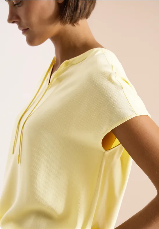 Seitliche Ansicht: Frau in hellgelbem Kurzarmshirt mit Bindeband am Halsauschnitt.