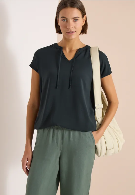 Frau in schwarzem Kurzarmshirt mit Kordelzug, grünen Leinenhosen und beiger Schultertasche.