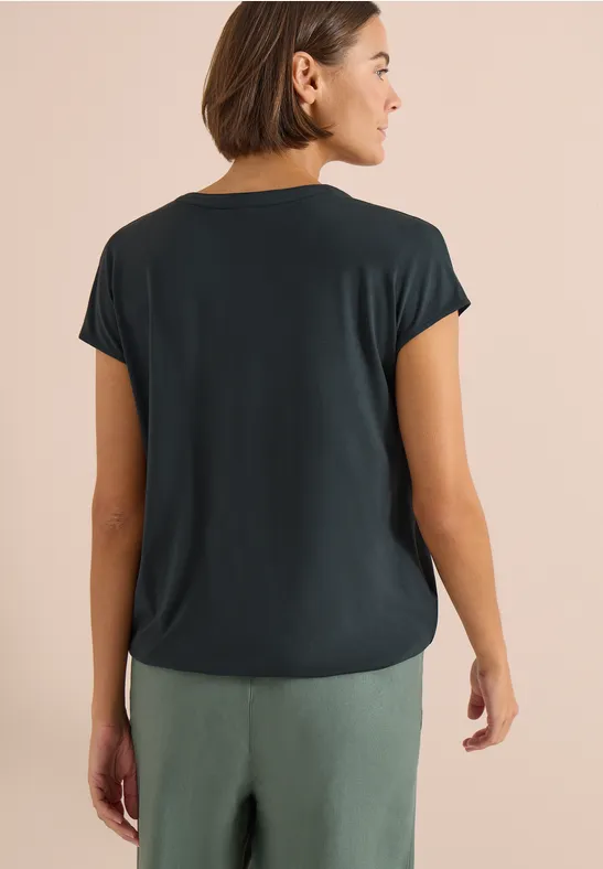 Frau von hinten in locker sitzendem dunkelgrauem T-Shirt und grüner Hose vor hellem Hintergrund.