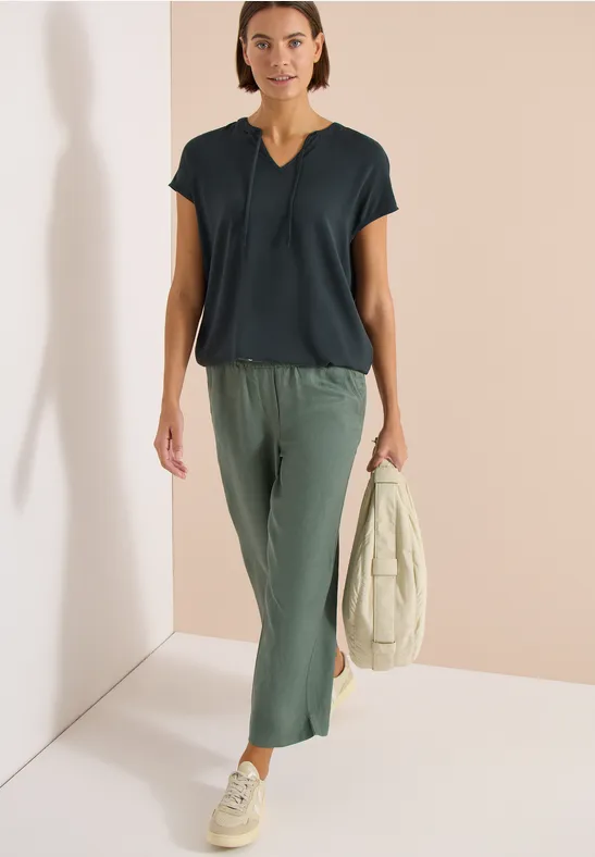 Frau in schwarzer Bluse, grüner Culotte-Hose und beigen Sneakern, hält beige Jacke, vor hellem Hintergrund.