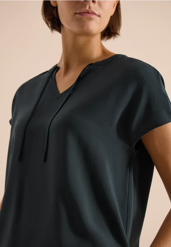 Eine dunkelgrüne, kurzärmlige Bluse mit V-Ausschnitt und dünnen Bändern am Halsansatz.