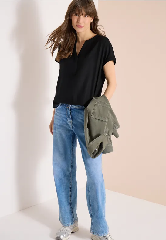 Eine Person in schwarzem T-Shirt, blauen Jeans und hält eine olivgrüne Jacke.