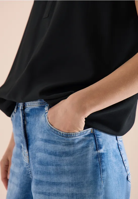 Person trägt schwarze Bluse und blaue Jeans, eine Hand in der Hosentasche.