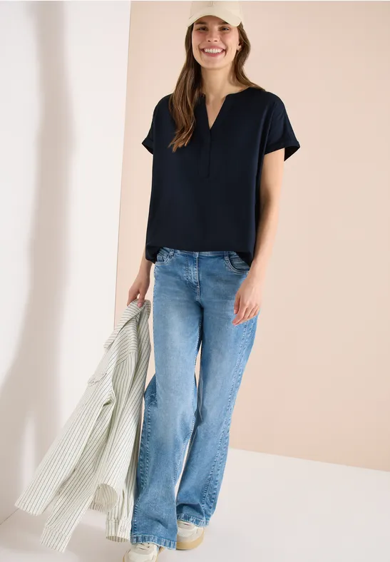 Frau in schwarzem V-Neck-Shirt, blauen Jeans, gelben Schuhen hält helle Jacke vor neutralem Hintergrund.
