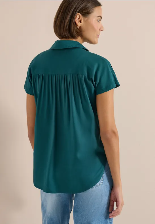 Rückansicht einer Frau in grünem Kurzarmbluse und hellblauer Jeans.