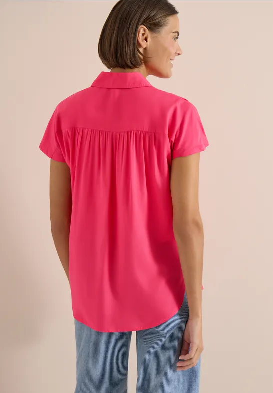 Frau mit Bobfrisur trägt von hinten ein pinkfarbenes Kurzarm-Hemd mit Kragen und Jeans.