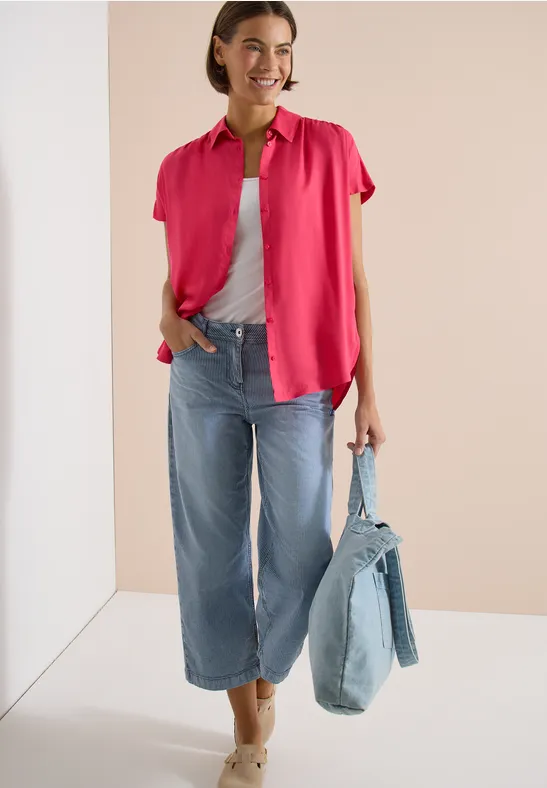Frau trägt pinke Kurzarmbluse über grauem Top, weite Jeans und hält eine Jeans-Tasche.
