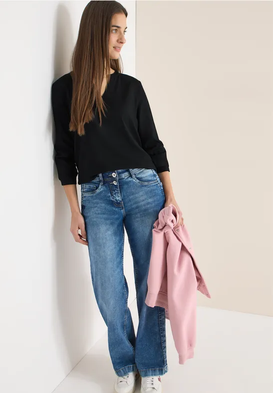 Frau in schwarzem Pulli und Jeans lehnt an einer Wand, hält eine rosa Jacke in der Hand.