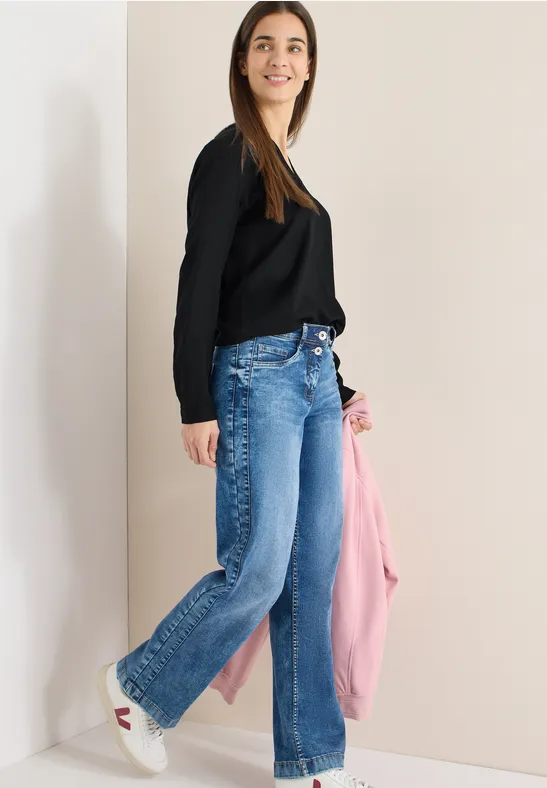 Frau trägt weite Jeans, schwarzen Pullover und hält eine rosafarbene Jacke vor beiger Wand.