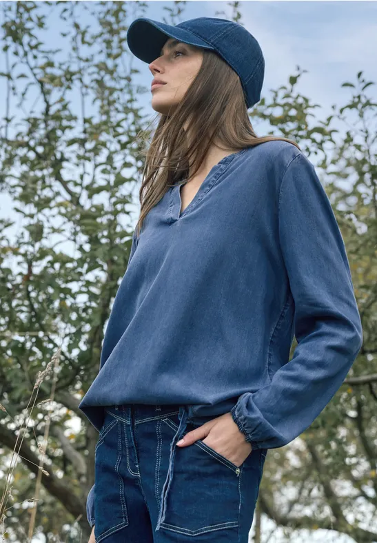 Eine Frau in blauer Bluse und Cap steht lässig vor einem blühenden Obstbaumbestand.