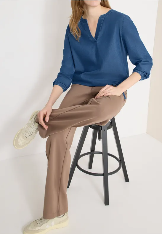 Frau sitzt auf schwarzem Hocker, trägt blaue Bluse, beige Hose und helle Sneaker vor weißem Hintergrund.