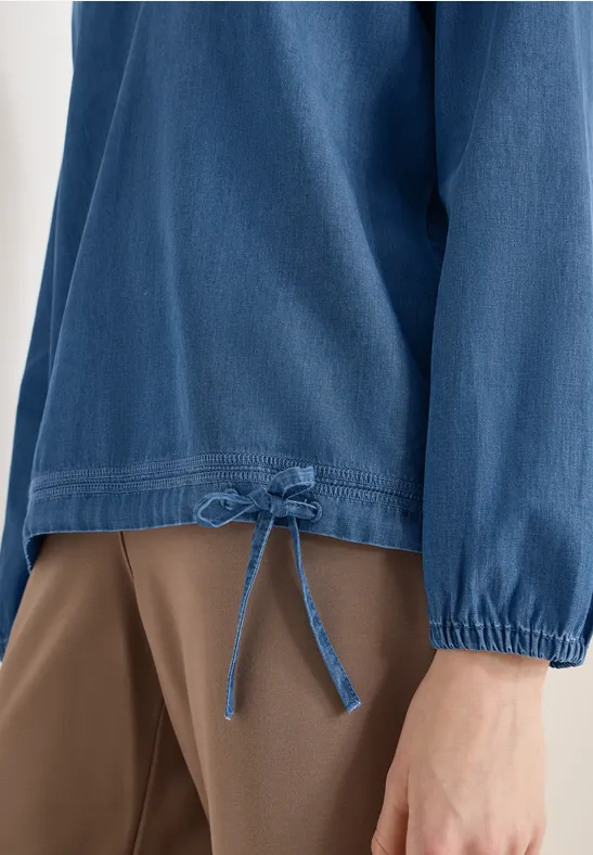 Detailaufnahme eines blauen Langarmshirts mit Tunnelzug und Schleife am Saum, kombiniert mit brauner Hose.
