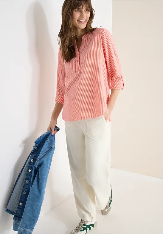 Frau in rosa Bluse, weiße Hose, hält Jeansjacke in der Hand, trägt grüne Slipper vor hellem Hintergrund.