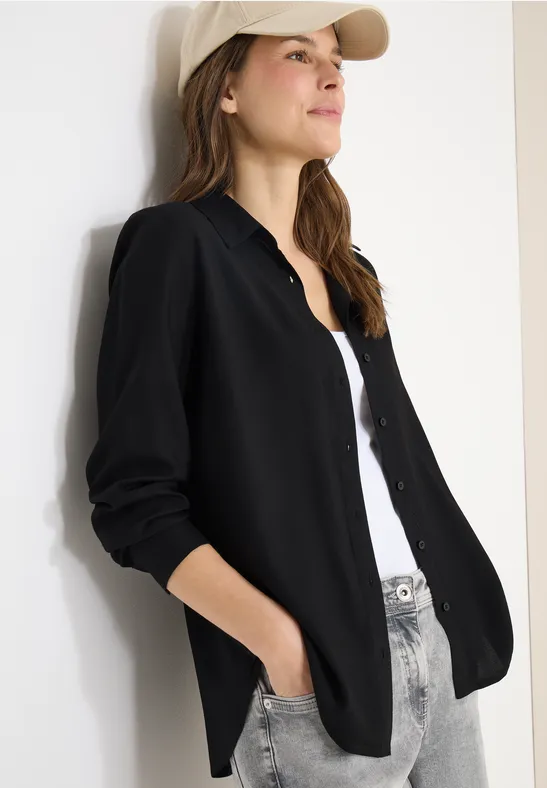 Person in schwarzer Hemdjacke, weißem Top, hellen Jeans und Cap lehnt an weißer Wand.