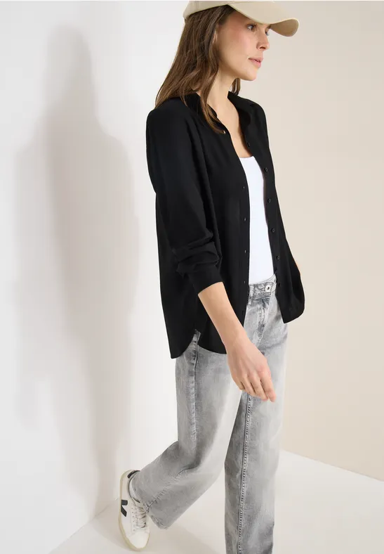 Frau in schwarzer Jacke, weißem Top und hellgrauen Jeans vor weißer Wand.