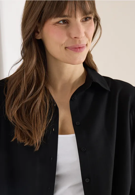 Frau mit langen braunen Haaren trägt ein offenes schwarzes Hemd über einem weißen Top.