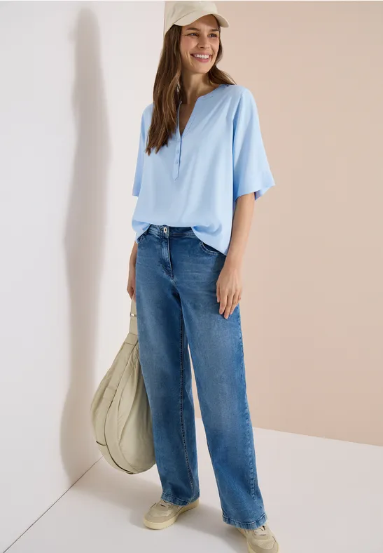 Person trägt hellblaues Oberteil, weite Jeans, helle Sneaker und große beige Tasche vor hellem Hintergrund.