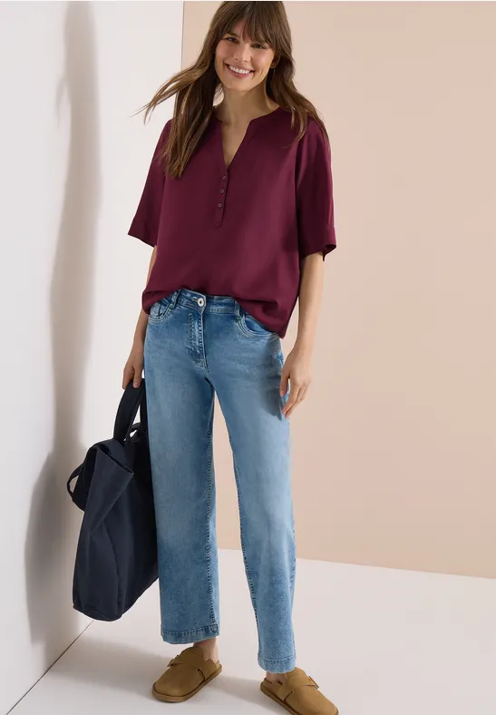 Frau steht vor hellem Hintergrund, trägt lila Bluse, gerade geschnittene blaue Jeans, braune Schuhe, hält große Tasche.