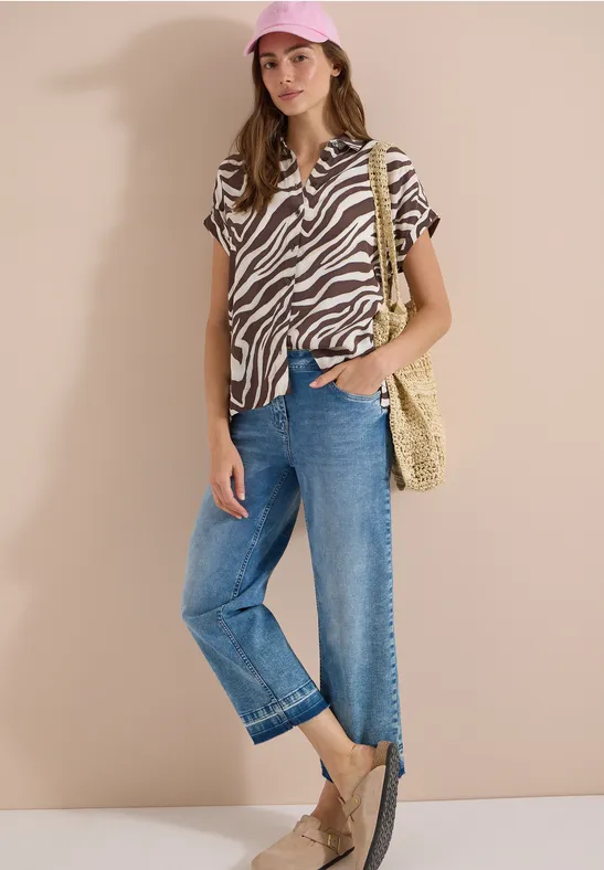 Femme en chemisier imprimé zèbre, jean ample, sac en paille, espadrilles et casquette devant un mur beige.