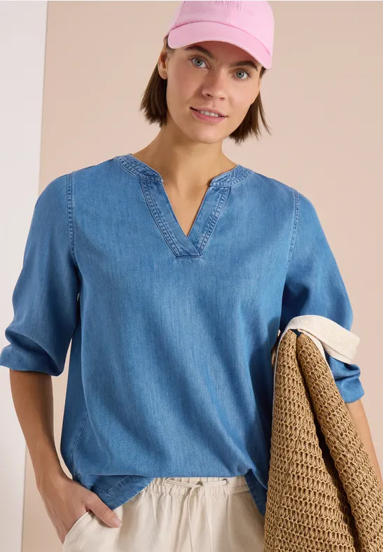 Femme portant une blouse en denim léger à col en V et manches 3/4, tenant un pull en maille.
