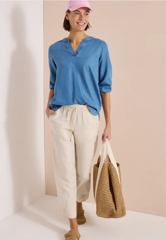 Femme en chemisier bleu, pantalon beige et sac en paille devant un mur bicolore.