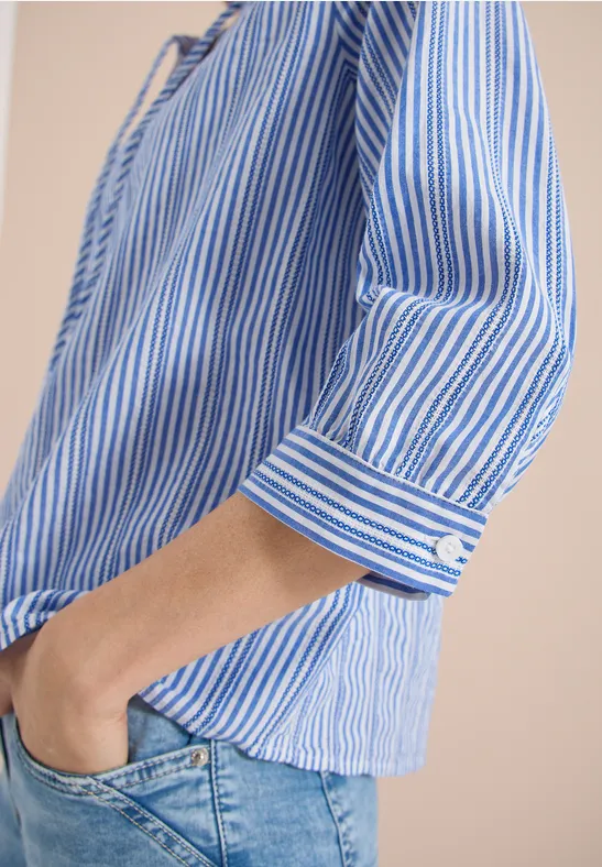 Torso einer Person in blau-weiß gestreifter Bluse mit hochgekrempelten Ärmeln und Jeans, Hand in Tasche.