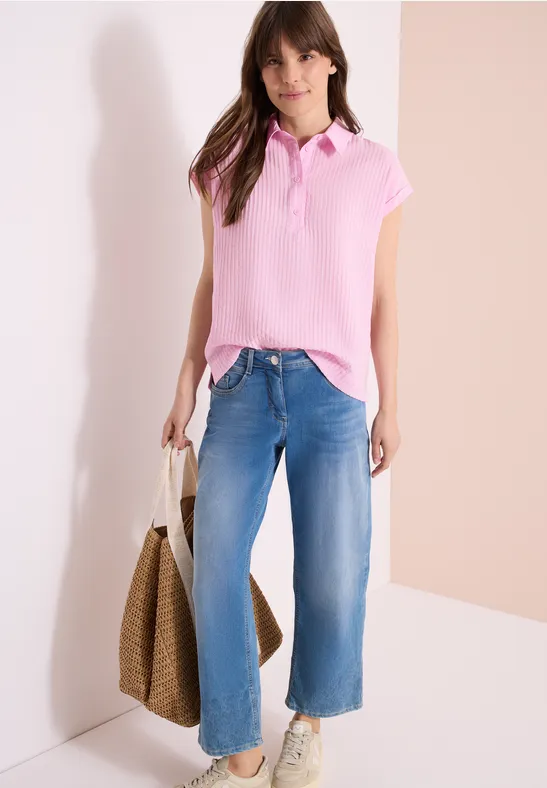Femme en polo rose, jean large bleu clair, sac en paille tressée, posant devant un mur clair.