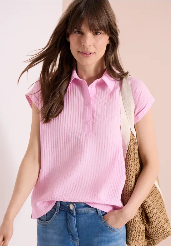 Jeune femme en polo rose à manches courtes et sac en paille, poignet dans la poche, ambiance décontractée.