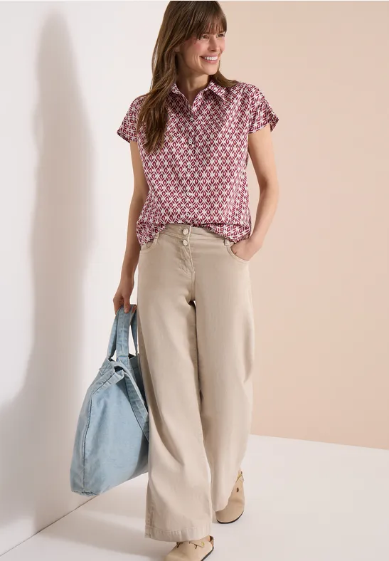 Frau trägt rosa gemustertes Oberteil, beige weite Hose und hält großen hellblauen Shopper.