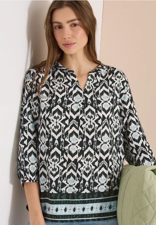 Eine Frau trägt eine schwarz-weiße, geometrisch gemusterte Bluse mit 3/4-Ärmeln und grünem Saum.