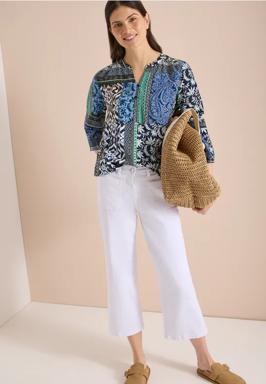 Frau trägt blaue gemusterte Bluse, weiße Schlagjeans, Strohtasche und braune Sandalen.