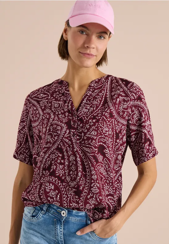 Frau in pinkem Cap und bordeaux Paisley-Bluse mit Jeans vor neutralem Hintergrund.