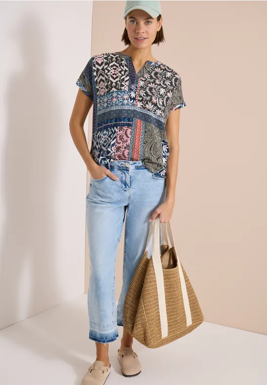 Frau trägt gemustertes Kurzarmshirt, hellblaue Jeans, Strohtasche und bequeme Espadrilles.