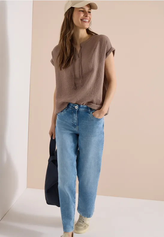 Frau mit locker sitzendem braunen Shirt, hellblauer Jeans, Sneakern, Cap und schwarzer Tasche.