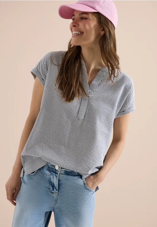 Junge Frau mit rosa Baseballcap, gestreifter Bluse und Jeans, lässig mit den Händen in den Taschen stehend.