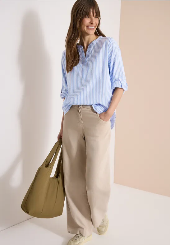 Eine Frau trägt hellblaues Hemd, beige weite Hose und olivgrüne Tasche vor neutralem Hintergrund.