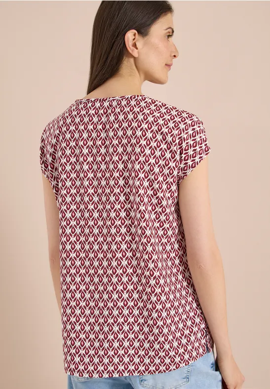 Frau von hinten in rotem, weißem Geometrie-Muster-Shirt vor beigem Hintergrund.