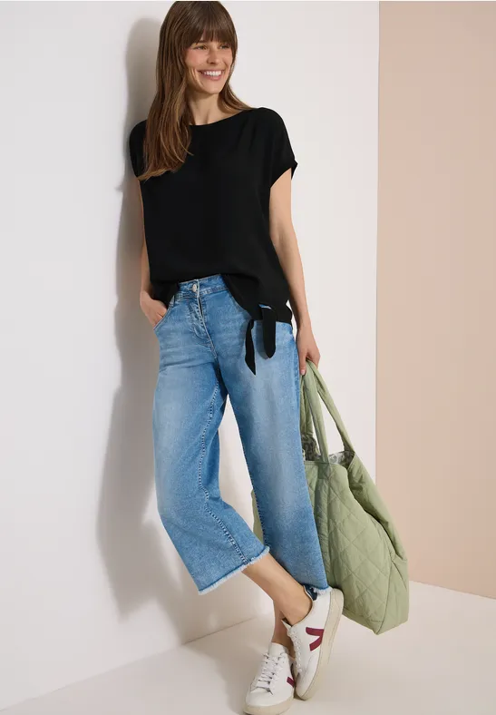 Femme en t-shirt noir, jean large court, baskets blanches à détail rouge et sac vert en tissu devant un mur clair.