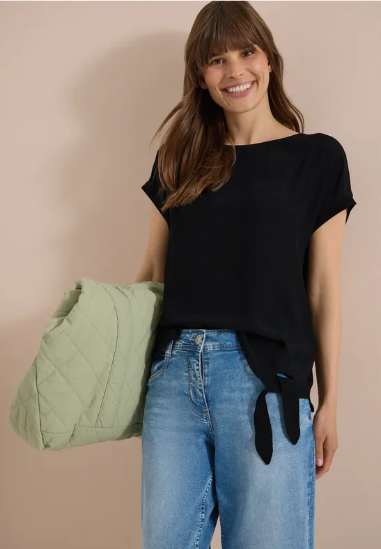 Femme en t-shirt noir à liens, jean bleu et grand sac matelassé vert.