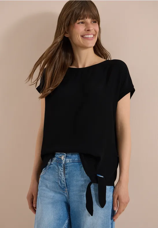Une femme porte un haut noir à manches courtes avec un nœud latéral et un jean bleu.