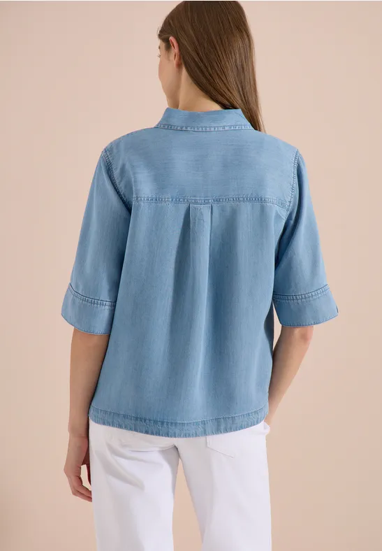 Rückansicht einer Frau in hellblauer Jeansbluse und weißer Hose.
