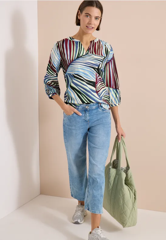 Frau mit bunter gestreifter Bluse, blauen Jeans und grünem Stoffbeutel vor heller Wand.