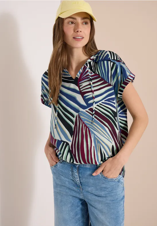 Femme portant un chemisier à motifs et un jean bleu sur fond clair.