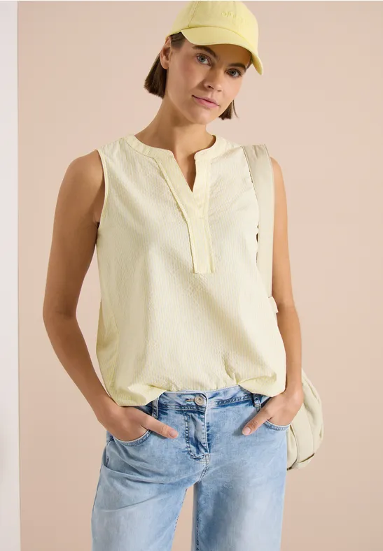 Personne portant un top sans manches jaune pâle et un jean bleu clair, mains dans les poches, fond rose.