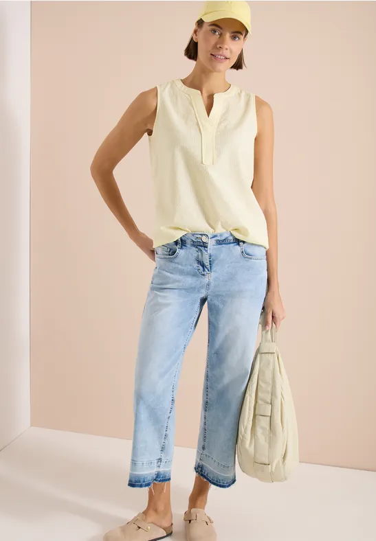 Femme en top sans manches crème, jean bleu clair et sac assorti devant fond neutre.