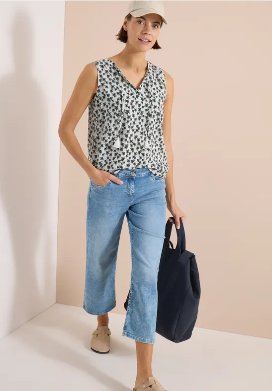 Frau in ärmellosem bedrucktem Top, weitem Jeans-Culotte, beigen Loafers, trägt schwarze Tasche vor hellem Hintergrund.
