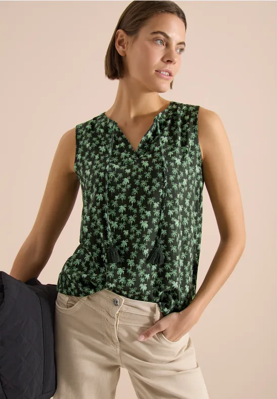 Ärmellose, grüne V-Neck-Bluse mit kleinem Blumenmuster, kombiniert mit beiger Hose vor hellem Hintergrund.