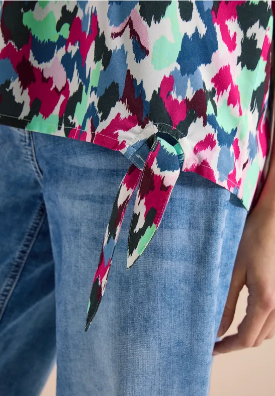 Farbintensives Top mit abstraktem Muster und Knoten am Saum, kombiniert mit Jeans.