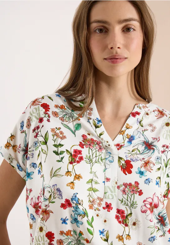 Frau trägt eine weiße, kurzärmlige Bluse mit buntem Blumenmuster.