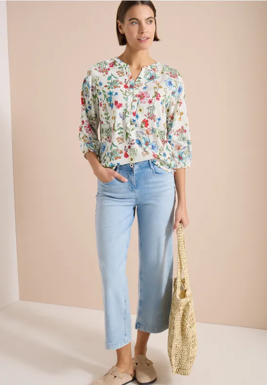 Frau mit geblümter Bluse, hellblauen weiten Jeans, Espadrilles und Strohtasche vor hellem Hintergrund.